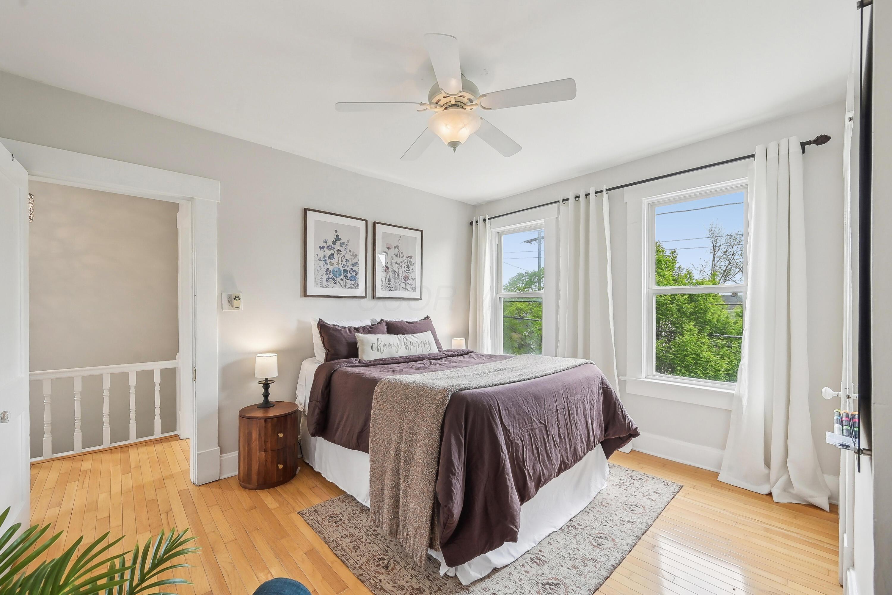Furnished bedroom with plum bedding, hardwood floors, a ceiling fan, and two curtained windows.