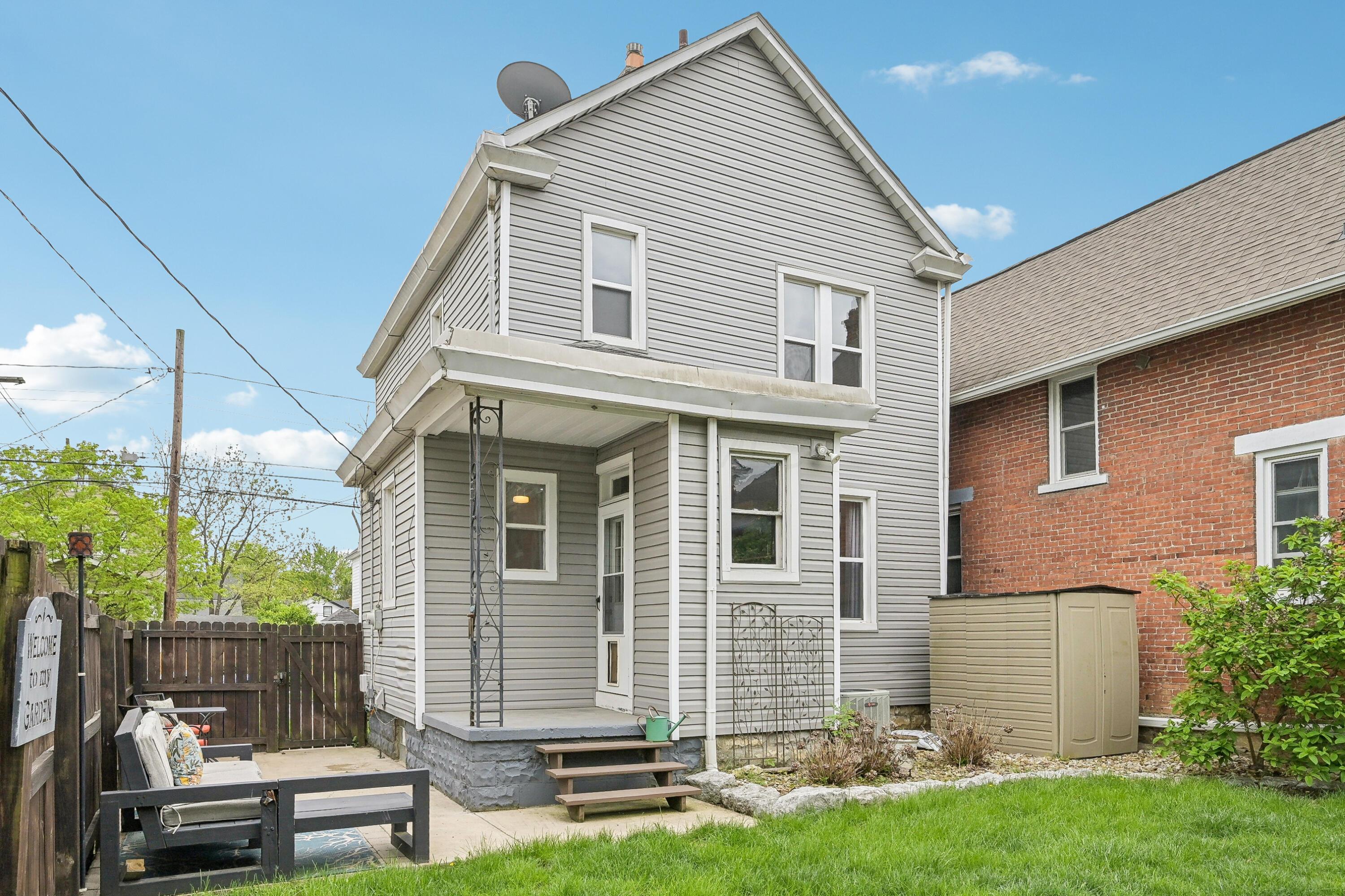 Gray two-story house with a covered rear porch, fenced patio seating area, and green lawn.