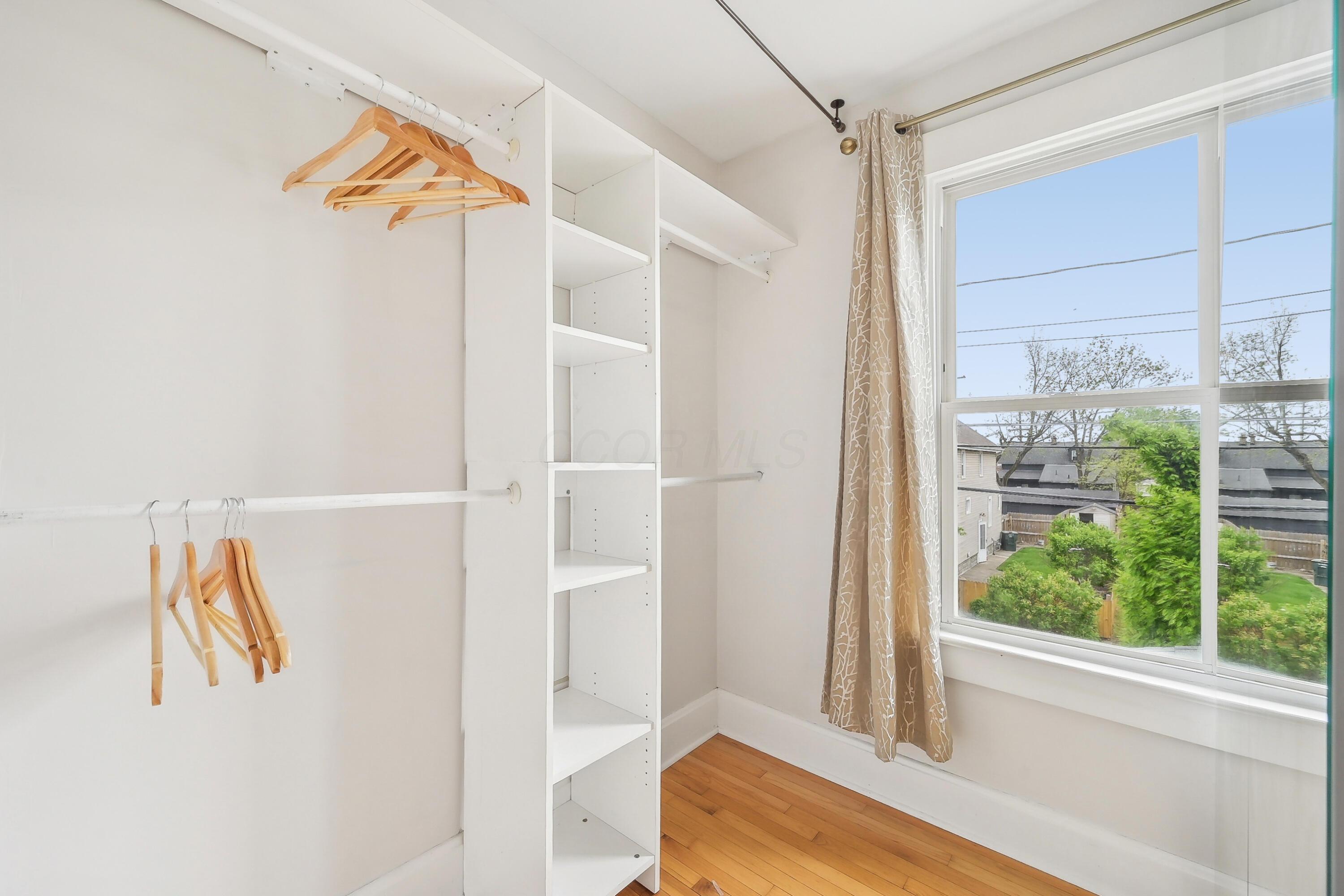 Walk-in closet with white built-in shelving, wooden hangers, hardwood floor, and a large backyard-facing window.