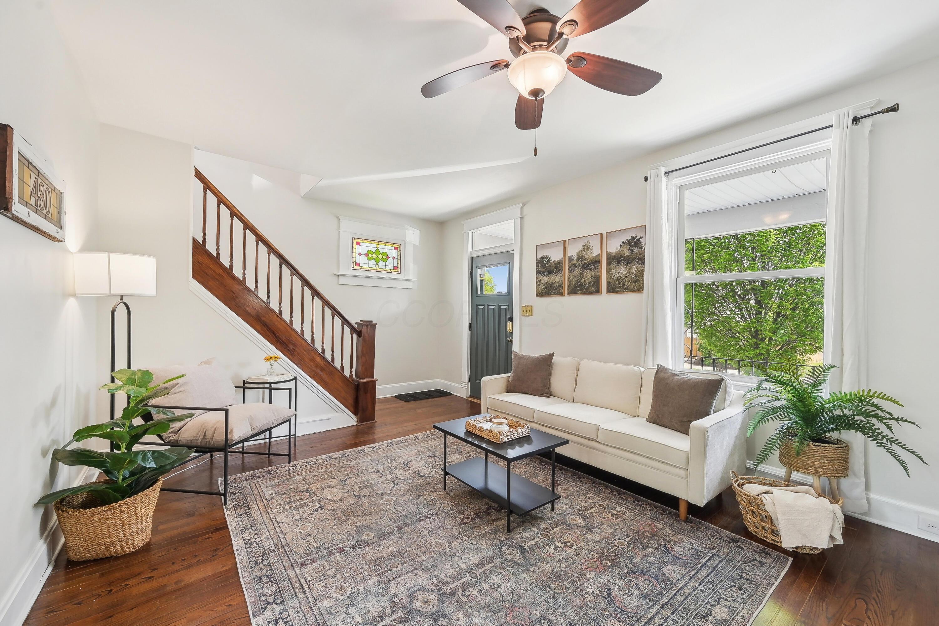 Staged living room with a wooden staircase, stained-glass window, and large window overlooking greenery.