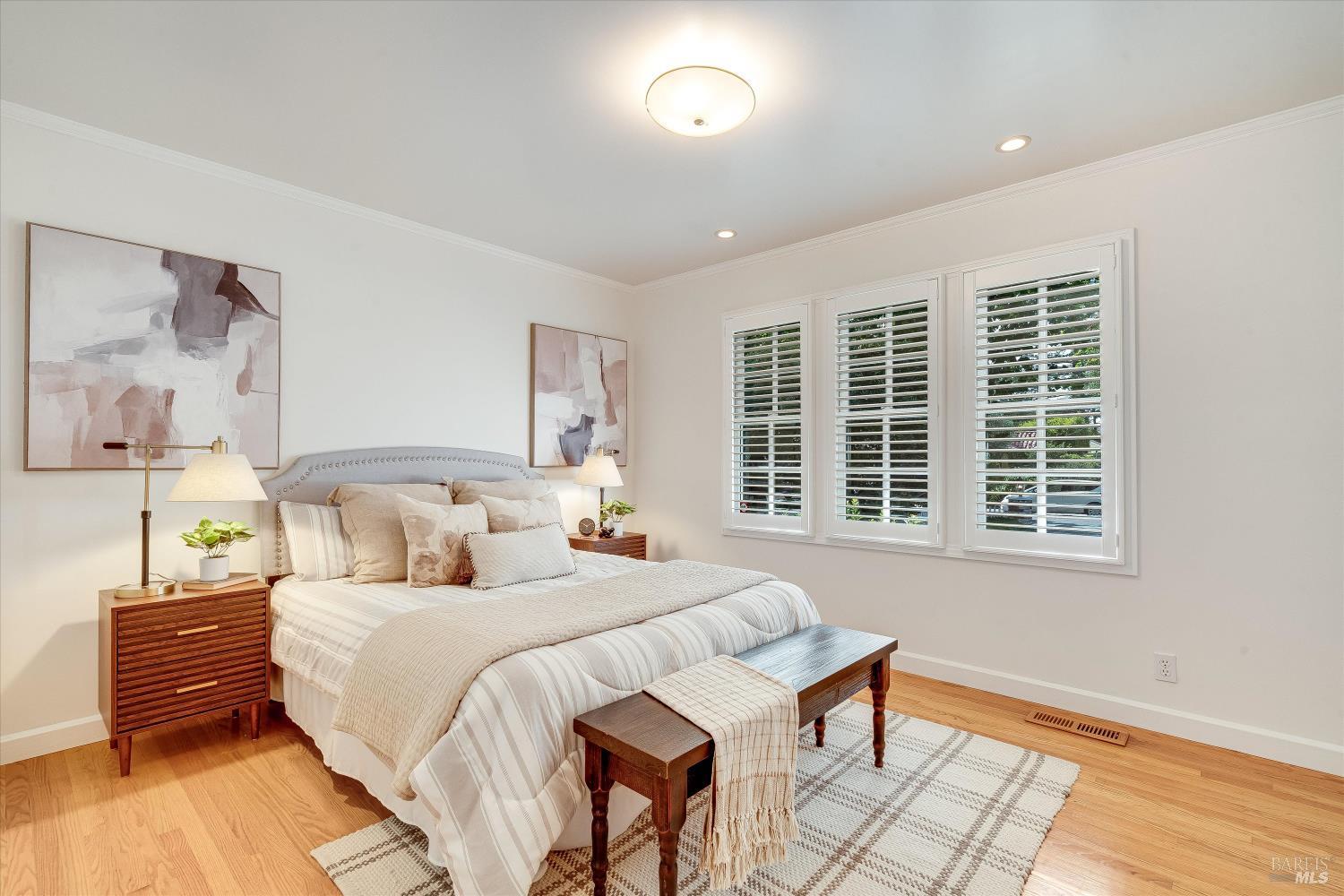 Furnished bedroom with pale striped bedding, wood nightstands, and three windows with plantation shutters.