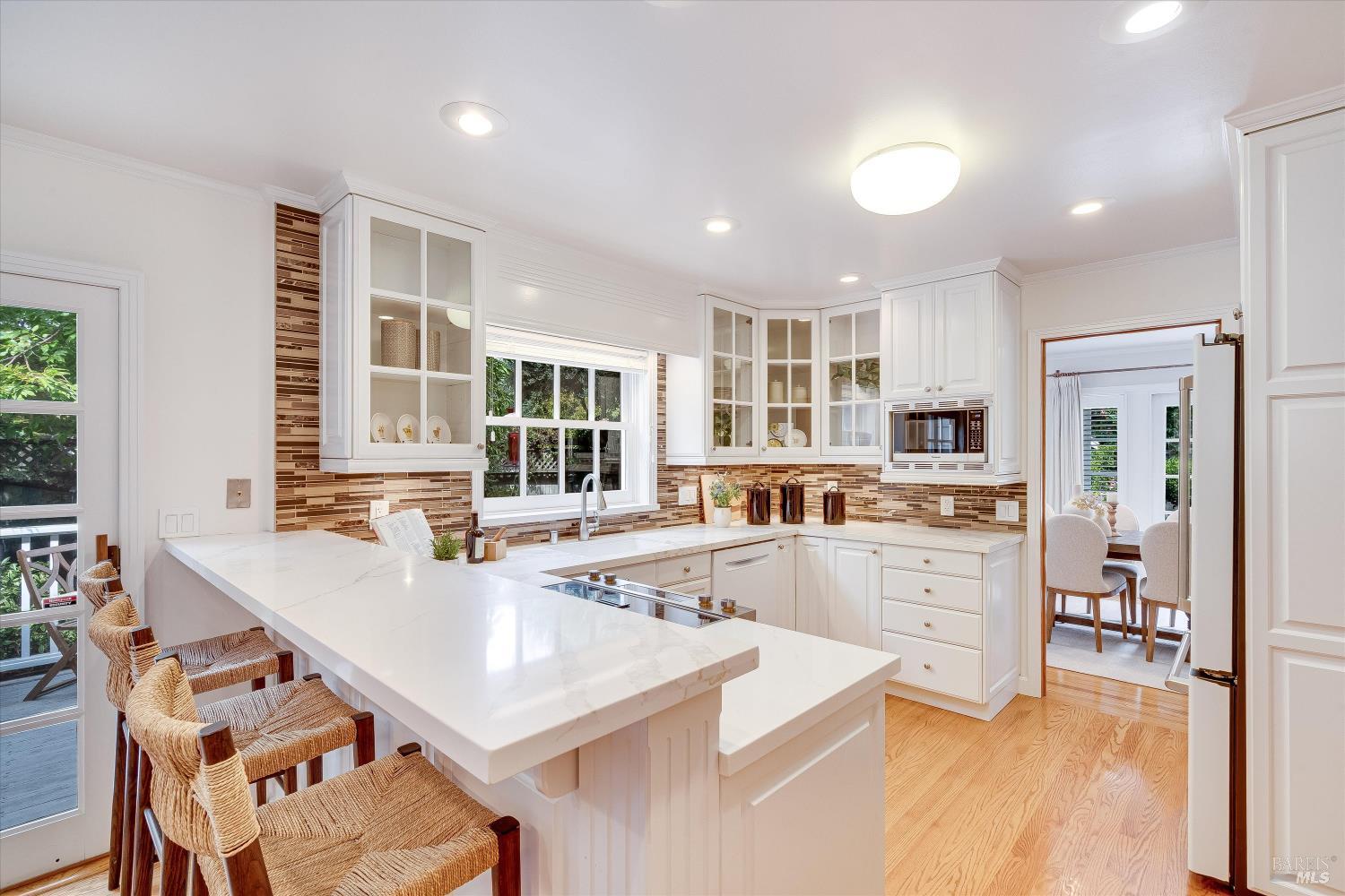 Bright white kitchen with a large stone island, woven stools, and mosaic tile backsplash.