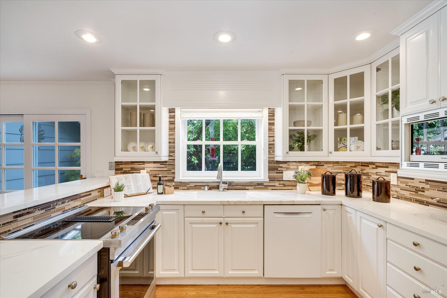 Bright white kitchen with glass-front cabinets, mosaic backsplash, and stainless-steel range.