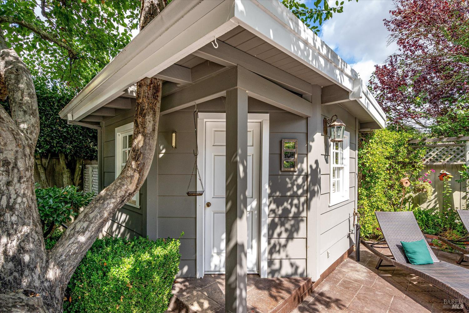 Gray structure with a covered entry beside a leafy garden patio and reclining chairs.