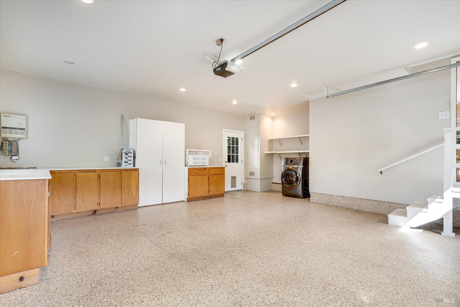 Spacious garage with speckled flooring, wooden cabinets, laundry machines, and stairs.