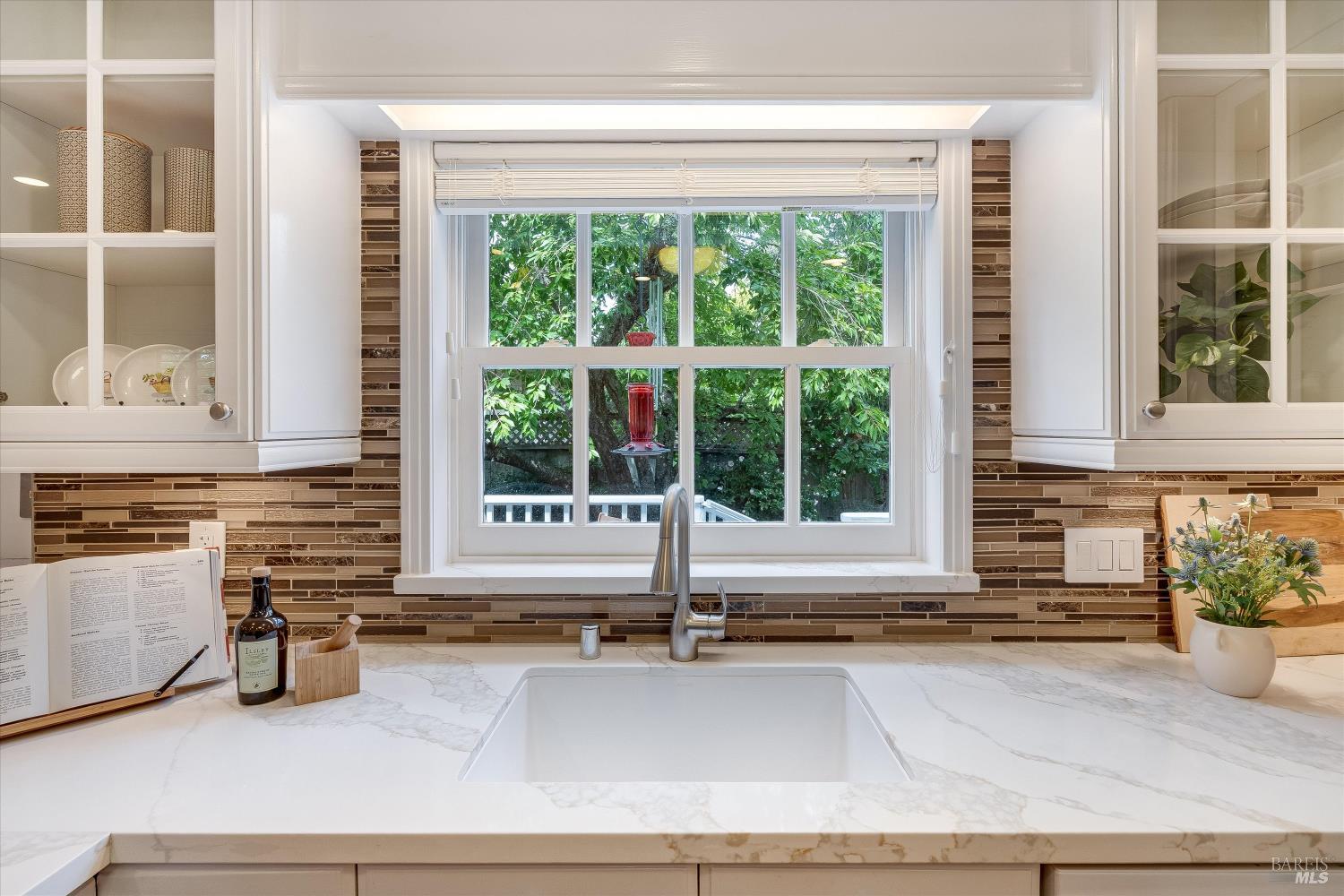 Kitchen sink beneath a large window, framed by white cabinets and a brown tile backsplash.