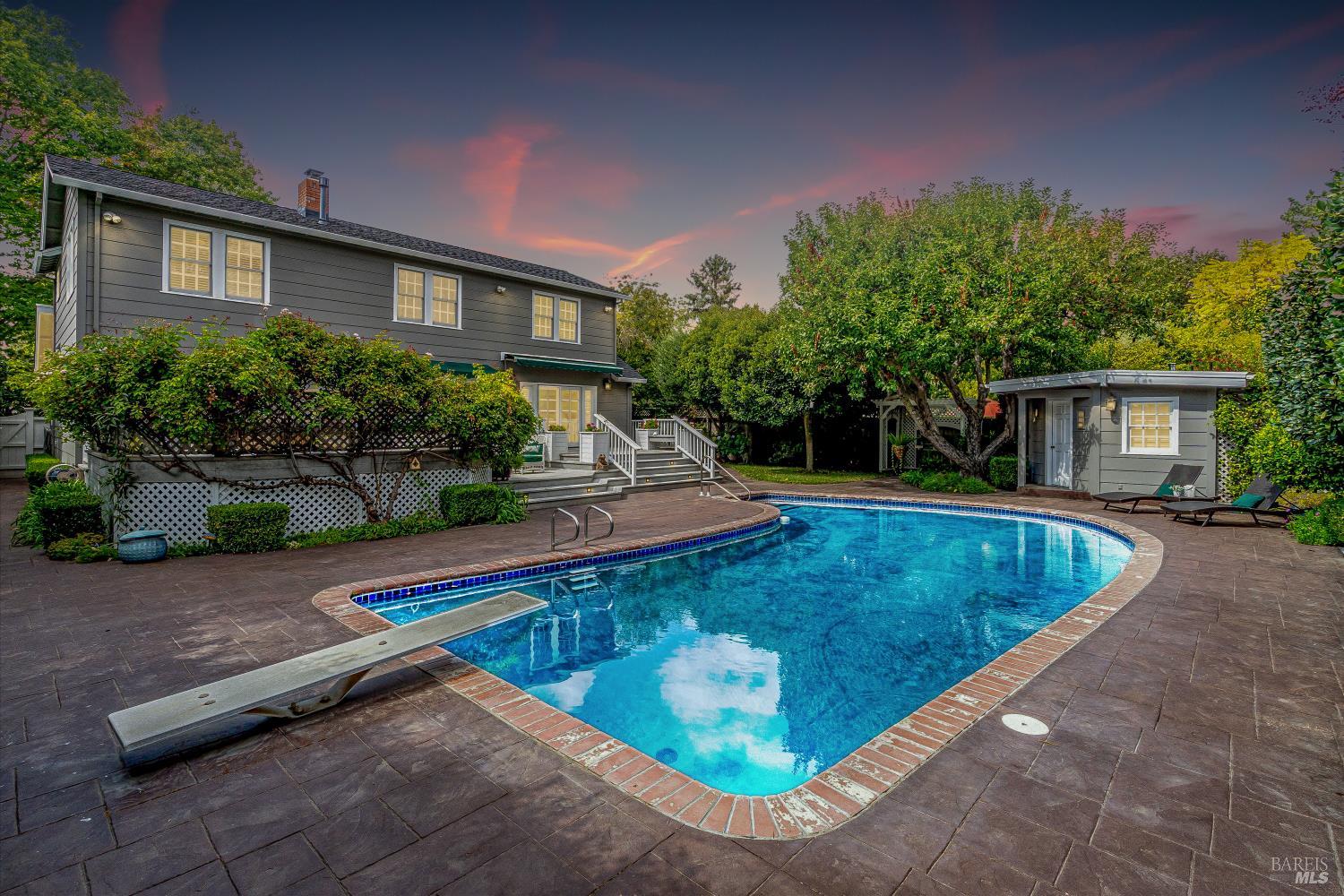 Backyard pool beside a gray two-story house, with a diving board and separate pool cabana.