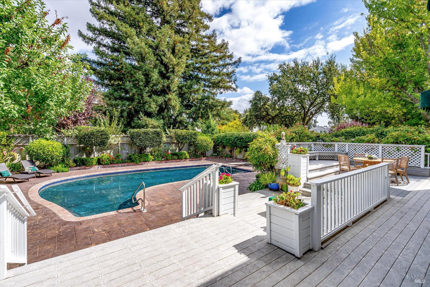 Backyard pool beside a white-railed deck, outdoor dining area, and mature leafy trees.