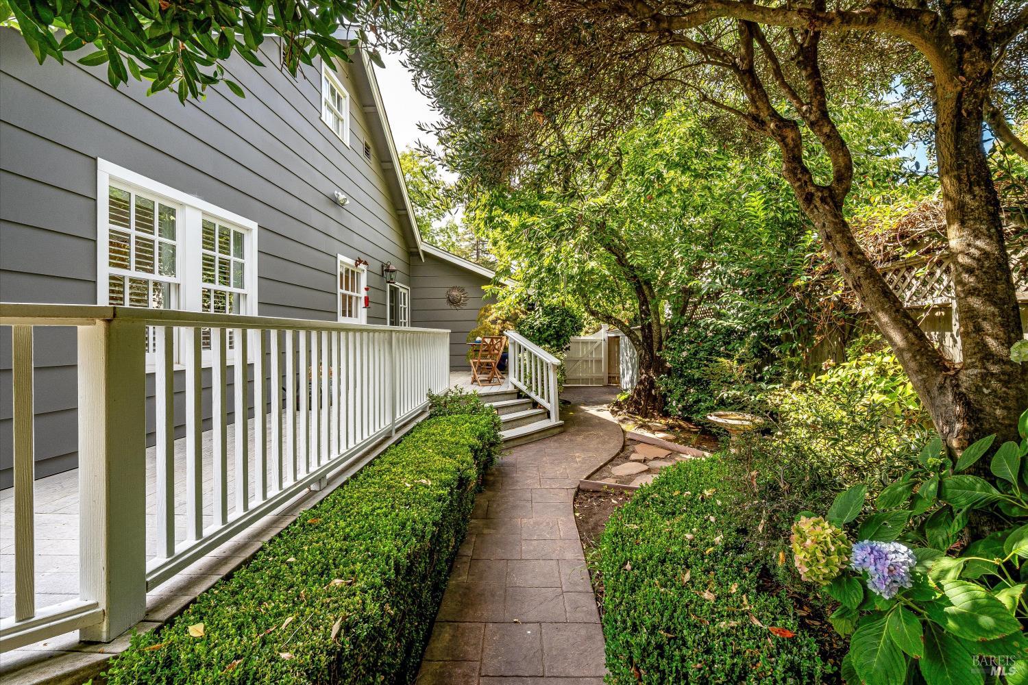 Gray home with a white-railed deck beside a curved garden path and mature trees.