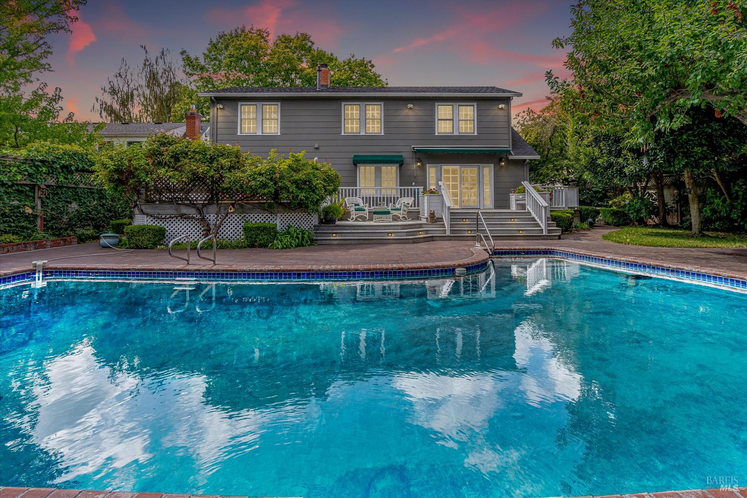 Two-story gray house behind a turquoise swimming pool, with a raised patio and mature landscaping.