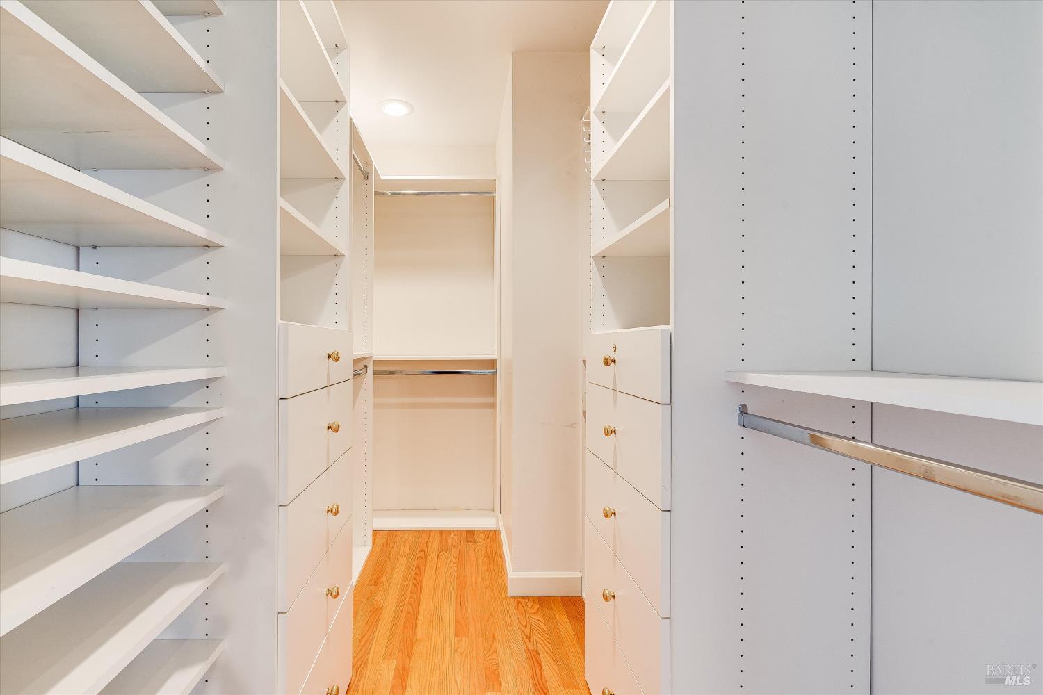 Walk-in closet with white built-in shelving, brass-knob drawers, and a warm wood floor.