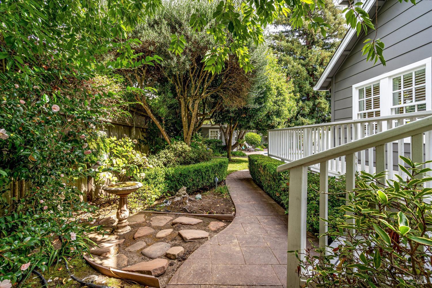 Landscaped garden path beside a gray home, with white porch railings and mature trees.