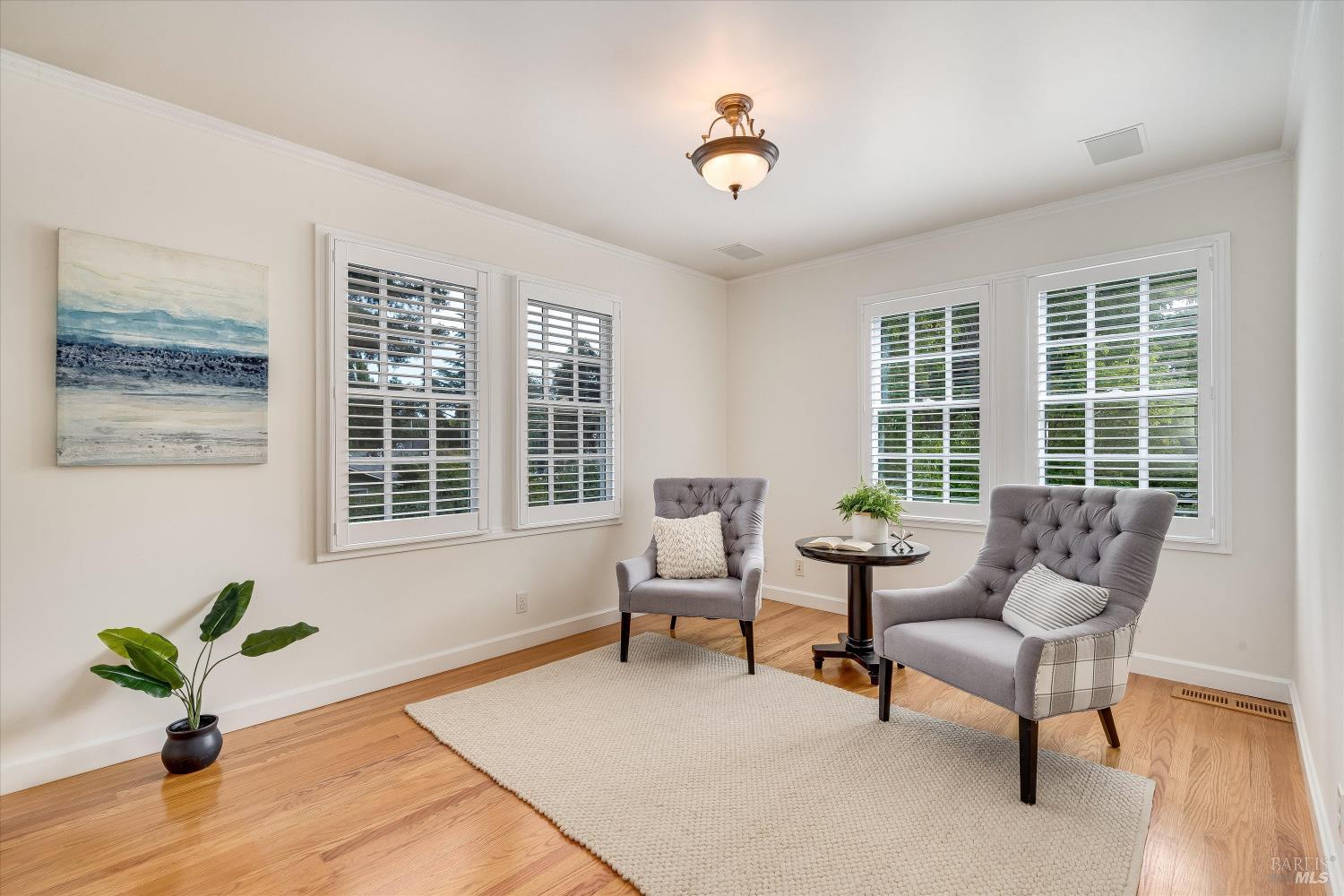 Staged sitting room with gray armchairs, a cream rug, hardwood floors, and large shuttered windows.