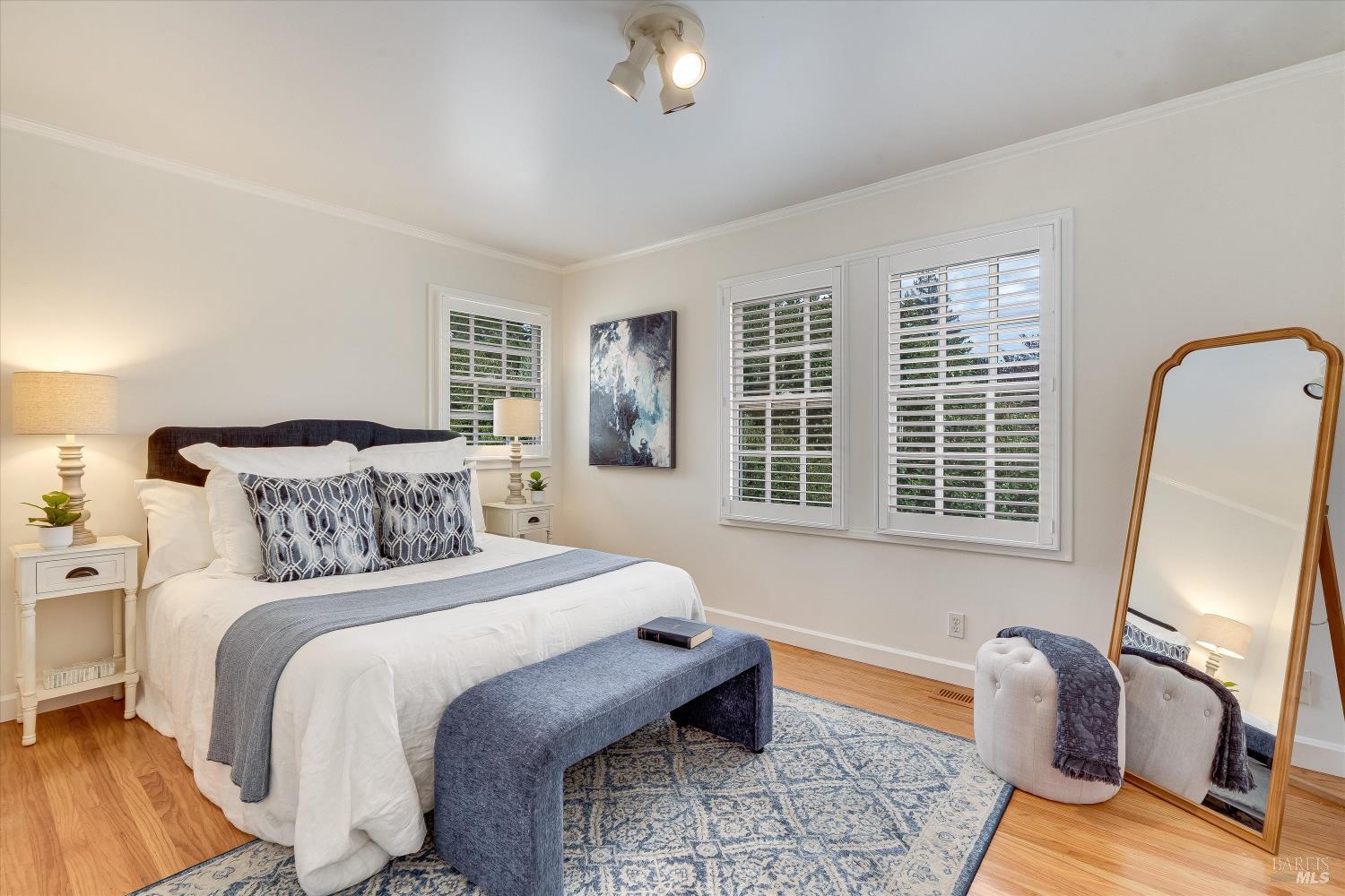Staged bedroom with a white bed, blue bench, patterned rug, and plantation-shuttered windows.