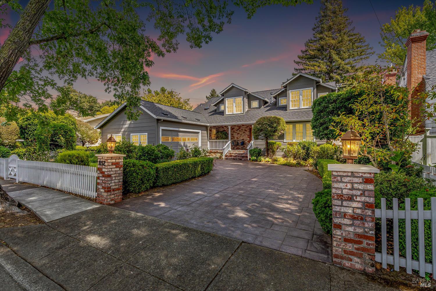 Gray two-story home with a broad paved driveway, white picket fence, and glowing entry lamps at dusk.