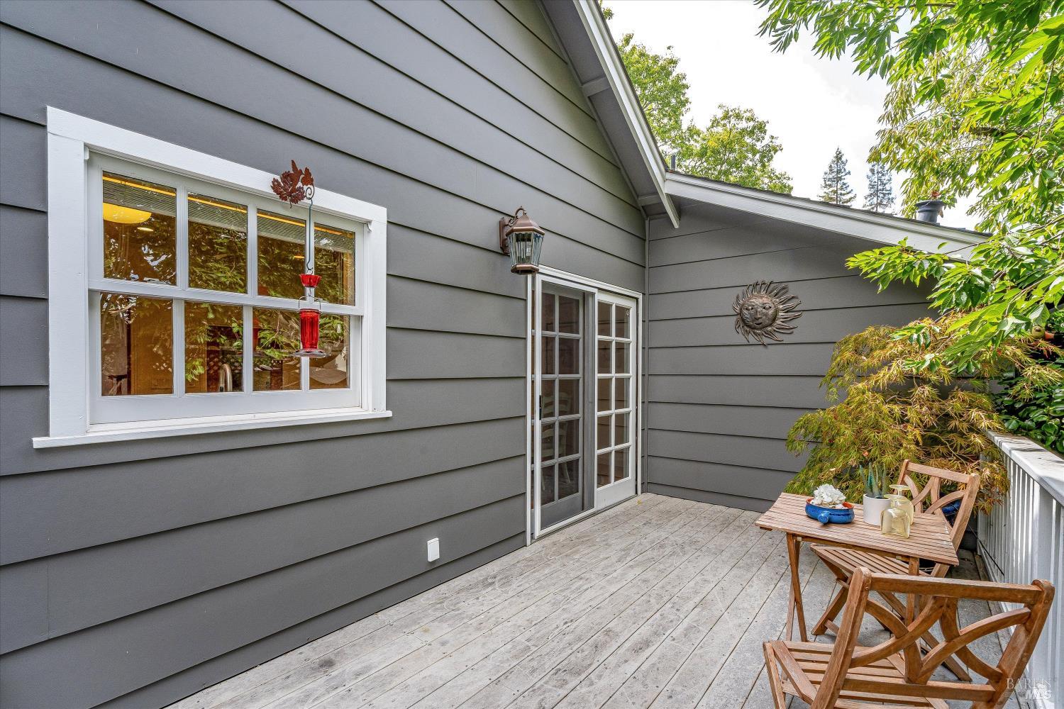 Wooden deck beside a gray home, furnished with a dining table and chairs among leafy plants.