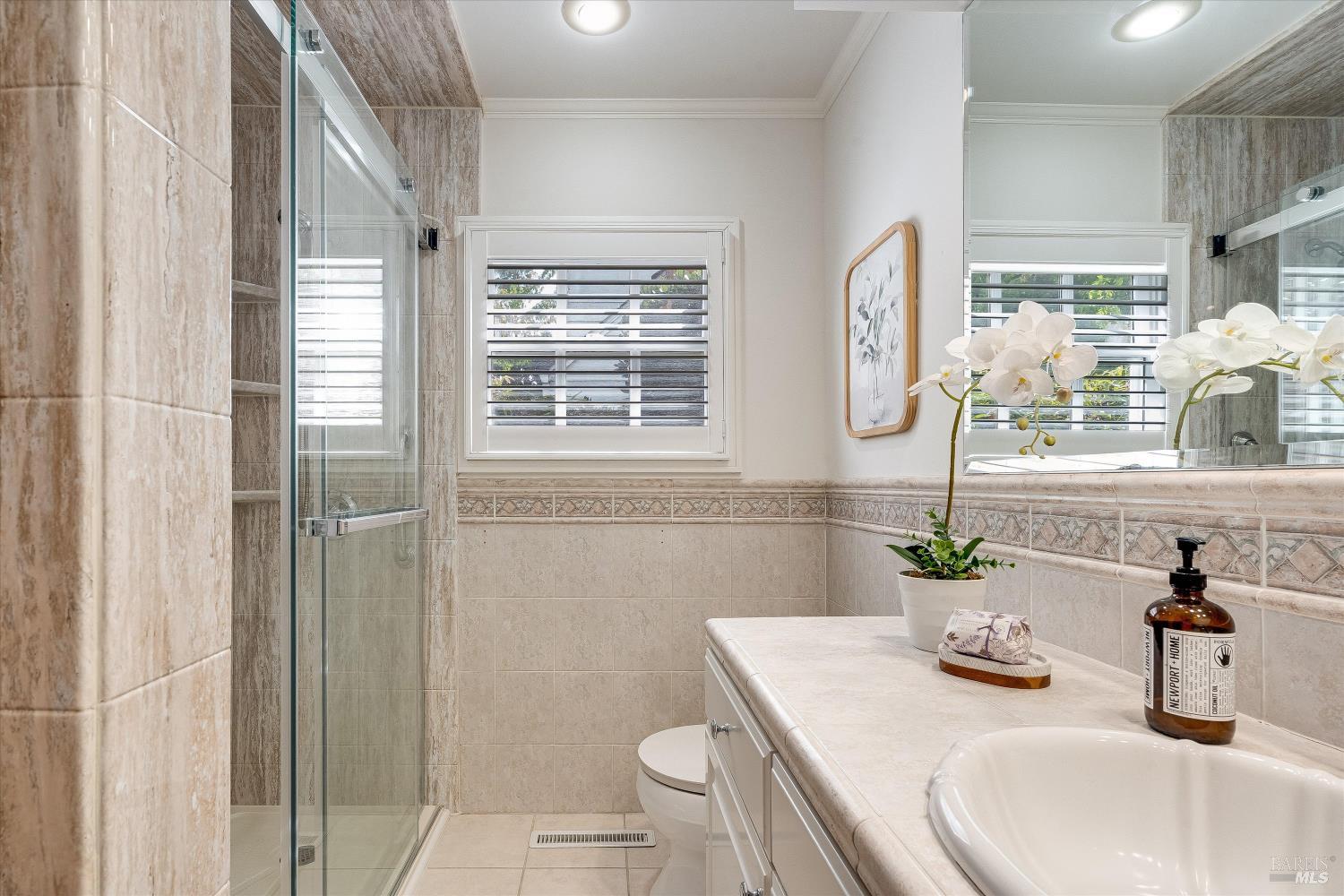 Bathroom with beige tiled shower, white vanity, shuttered window, and orchid.