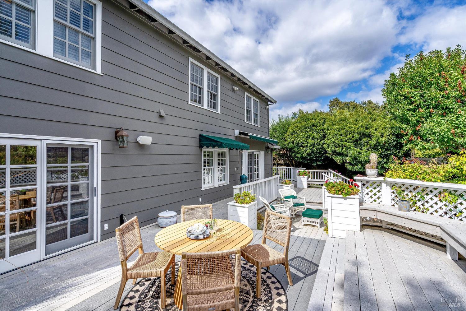 Furnished deck beside a gray two-story home, with outdoor dining furniture and flower planters.
