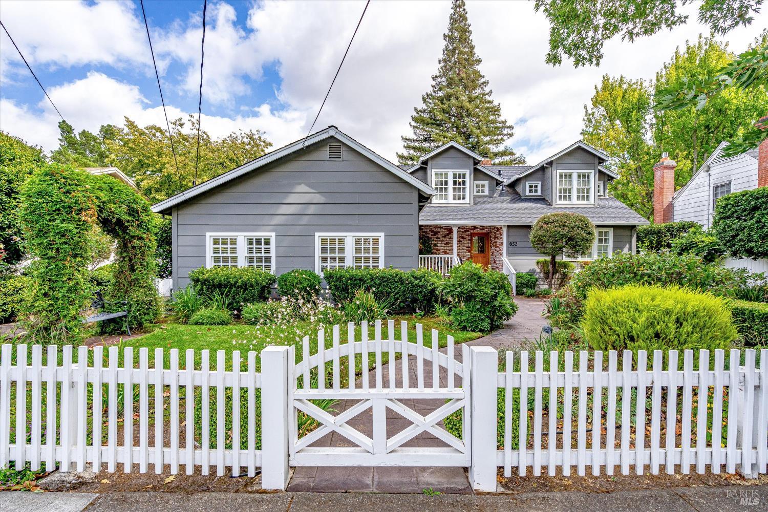 Gray two-story house with dormer windows, brick entry, and a white picket fence around its garden.