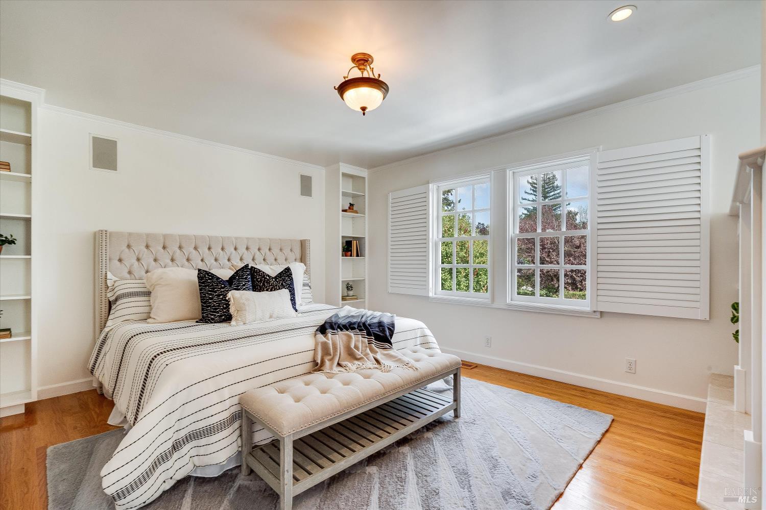 Staged bedroom with a tufted bed, cushioned bench, built-in shelves, and shuttered windows.