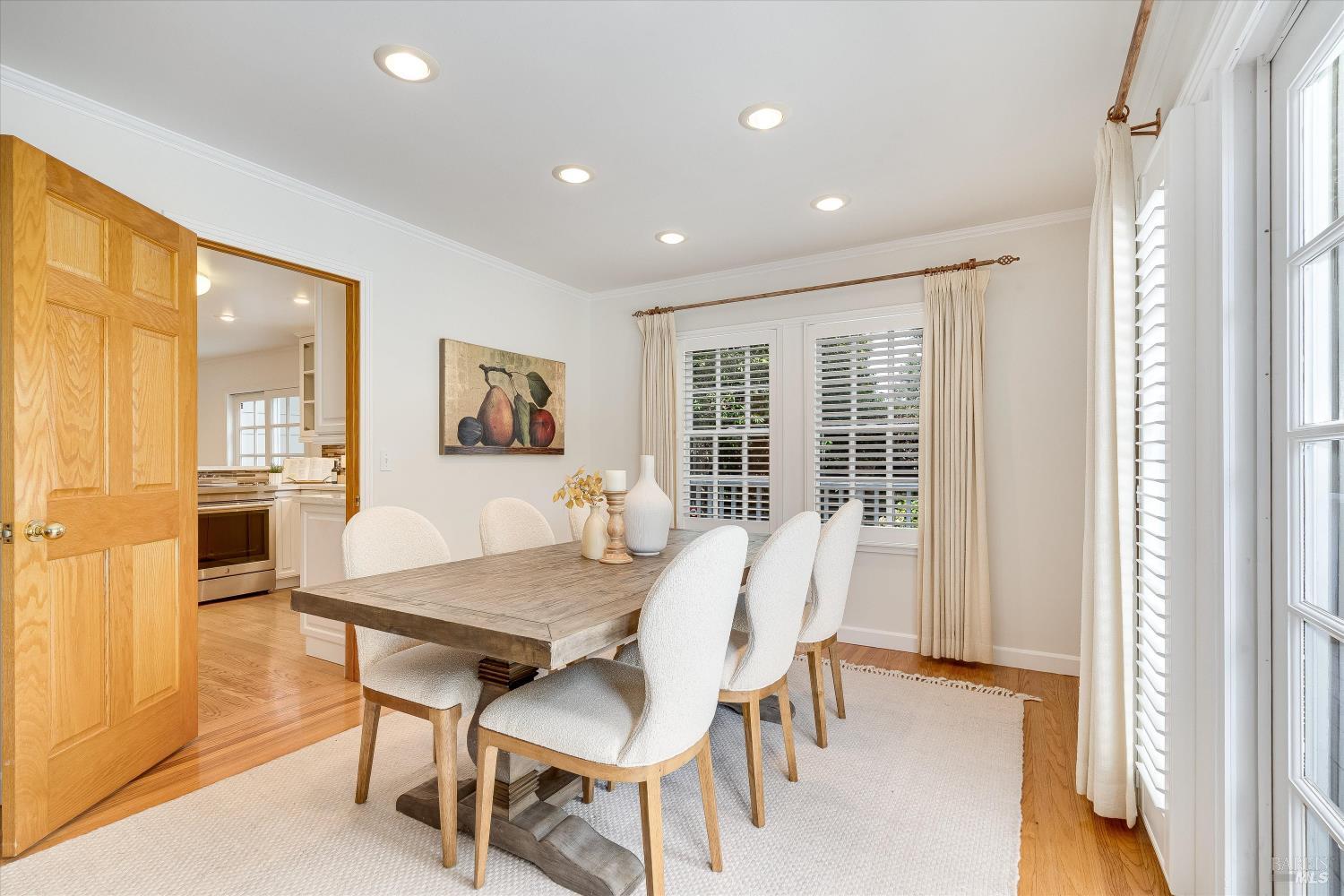 Dining room with a wood table, white chairs, and tall windows with plantation shutters.