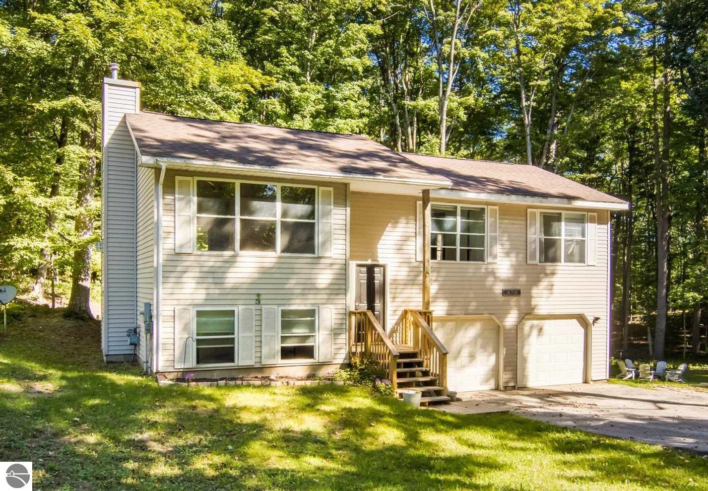 Beige split-level house with a two-car garage, front steps, and a wooded lawn.