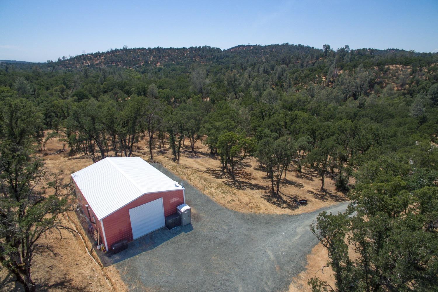 Aerial view of a red outbuilding with a white metal roof amid wooded acreage and a gravel drive.