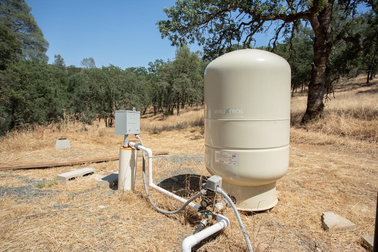 Well pressure tank and pump equipment with exposed pipes in a dry, oak-dotted clearing.