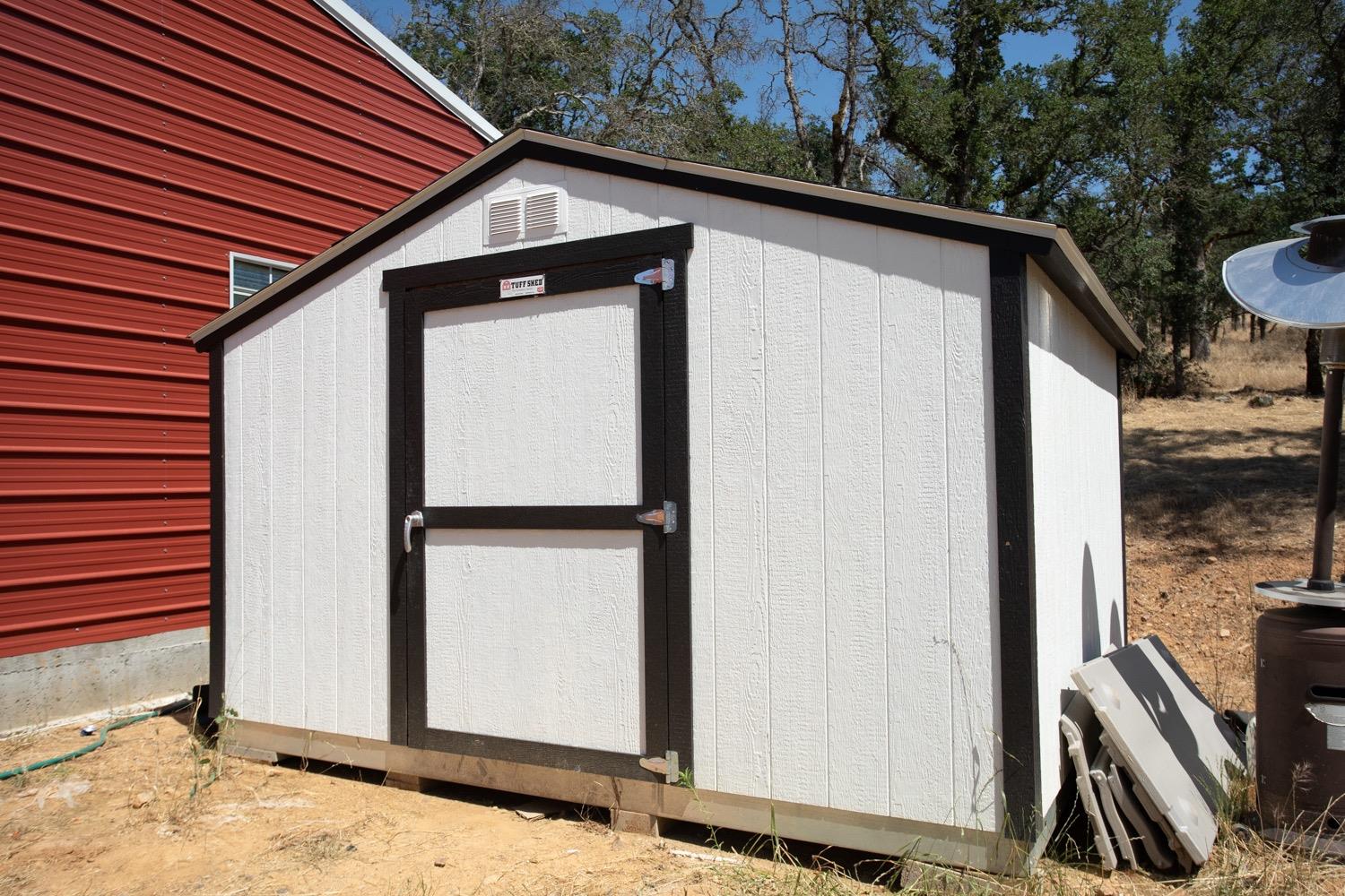 White storage shed with dark trim and a single door beside a red metal outbuilding.