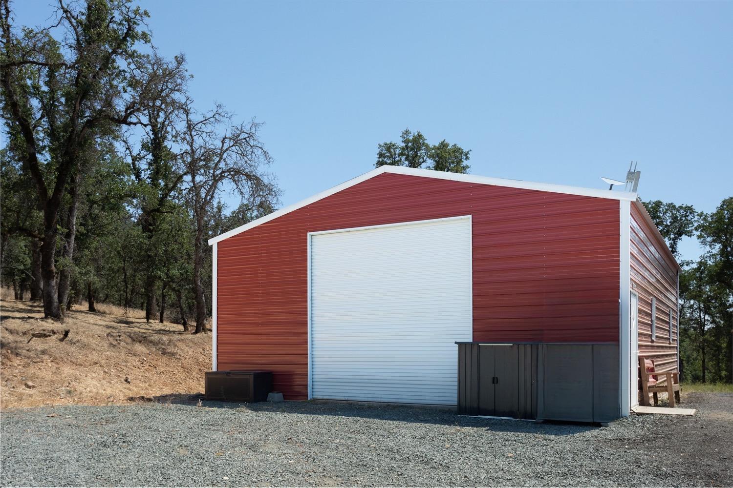 Red metal outbuilding with a wide white roll-up garage door in a gravel clearing.
