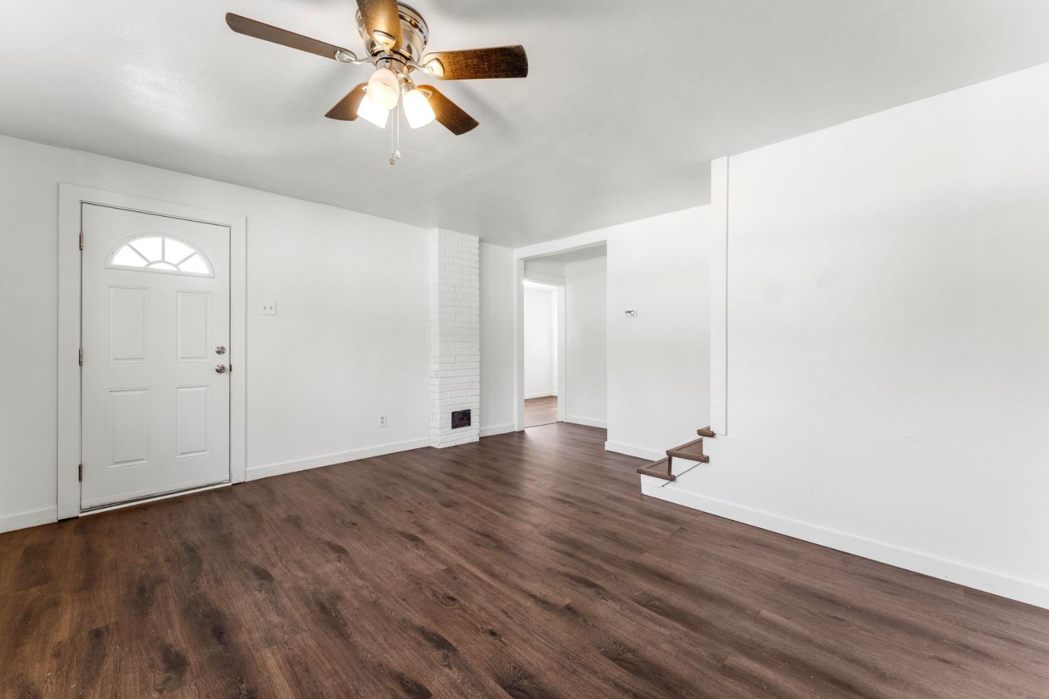 Vacant living room with dark flooring, a white brick fireplace, ceiling fan, and front door.