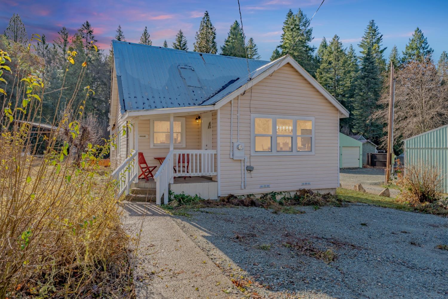 Beige house with a steep blue metal roof, white porch railings, and a gravel driveway at dusk.