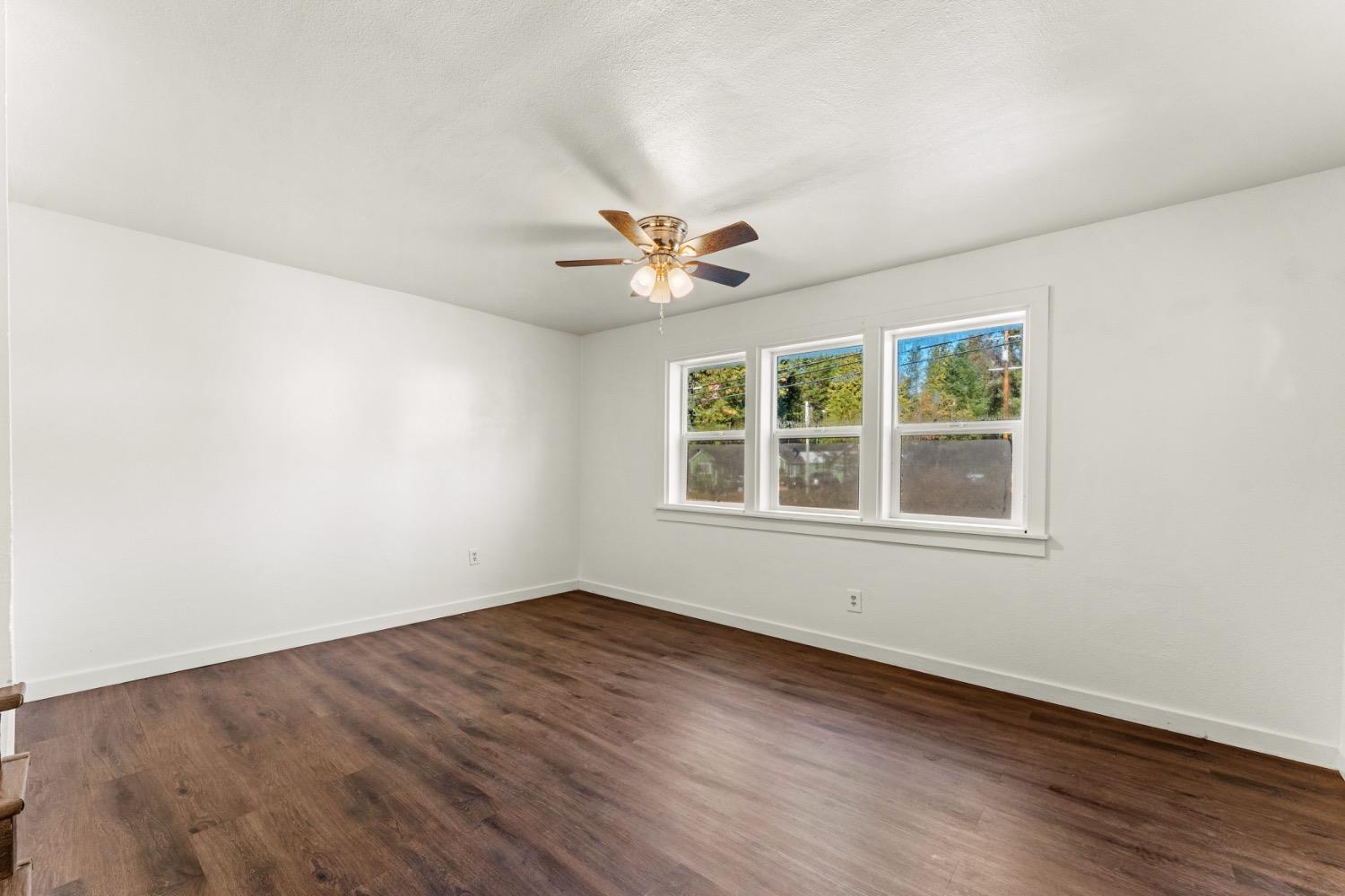 Empty room with dark wood-look floors, three windows, and a ceiling fan with lights.