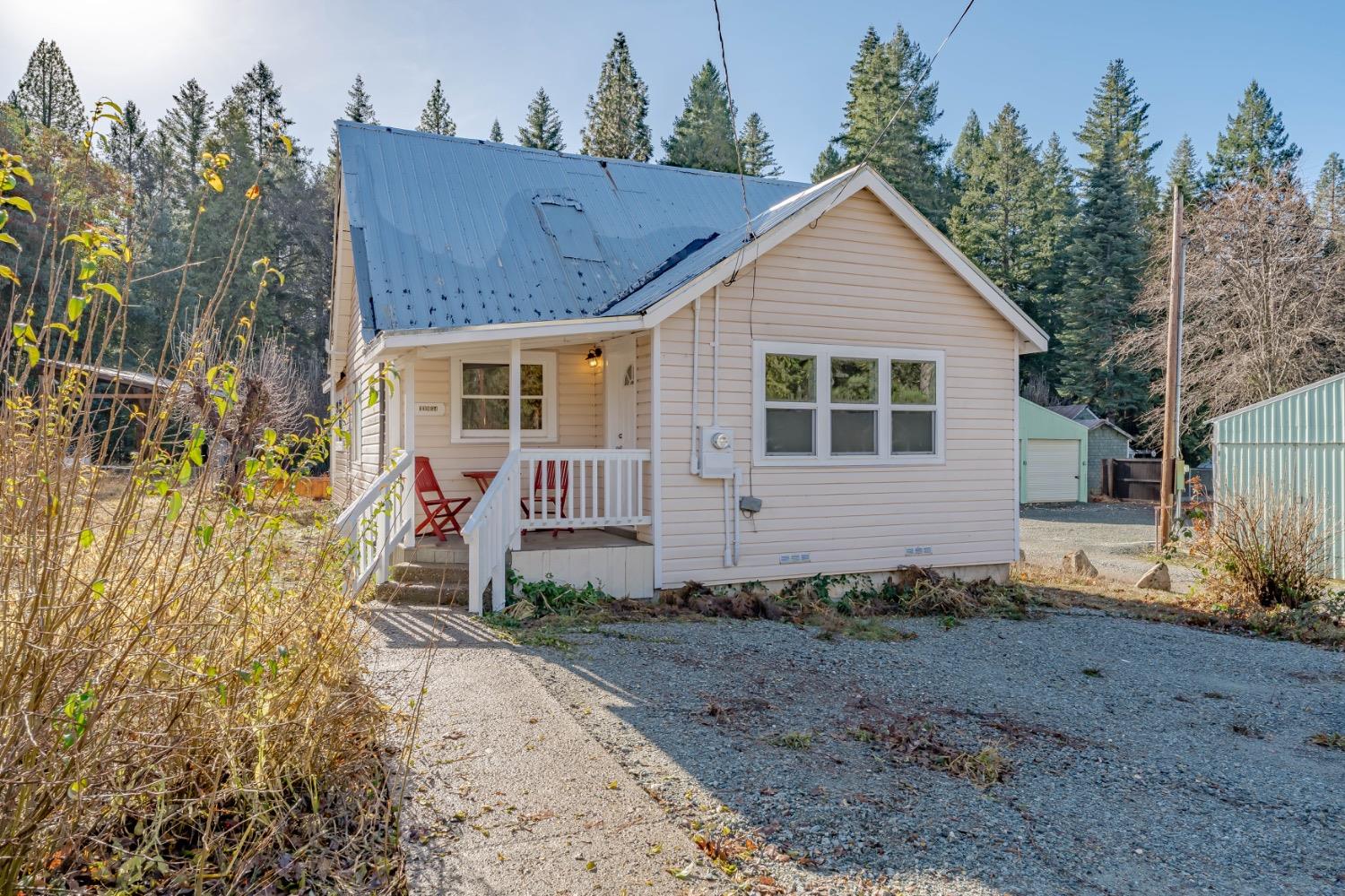 Small light-colored house with a blue metal roof, white porch railing, and red chairs amid evergreen trees.