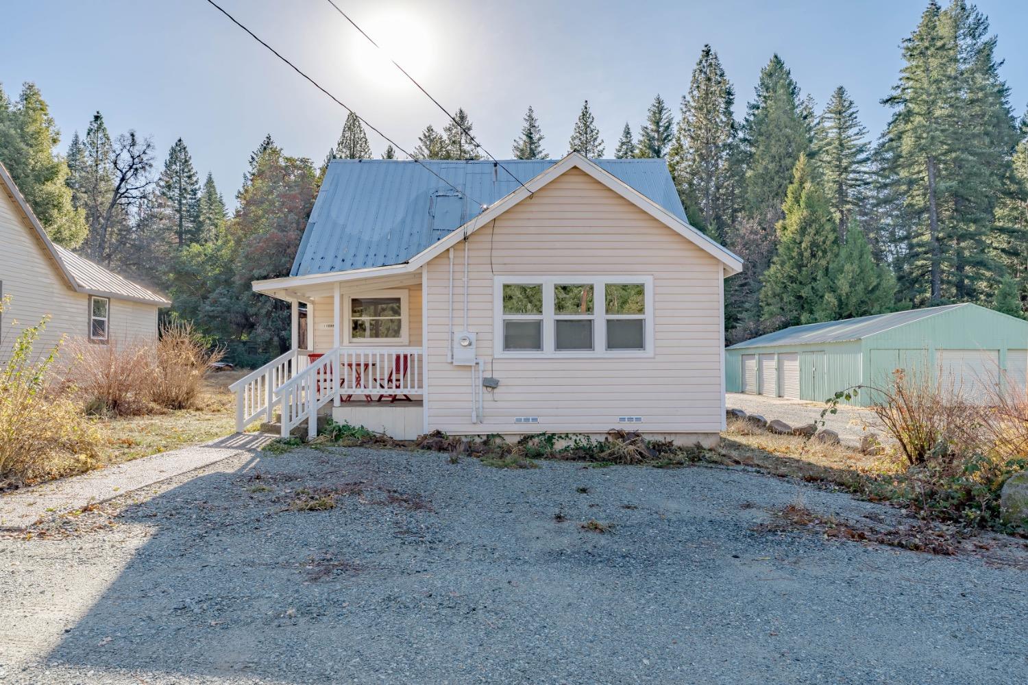 Beige house with a blue metal roof, white-railed entry ramp, gravel driveway, and wooded surroundings.