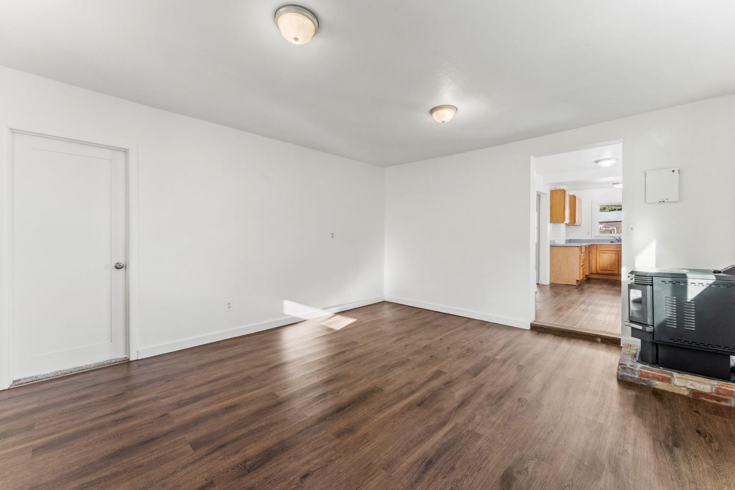 Vacant room with brown wood-look floors, a black stove on a brick hearth, and the kitchen visible through a doorway.