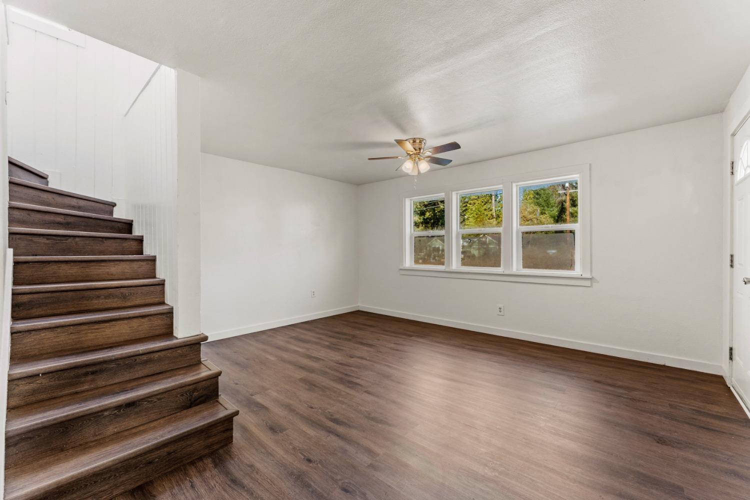 Vacant room with dark wood floors, a staircase, three windows, and a ceiling fan.