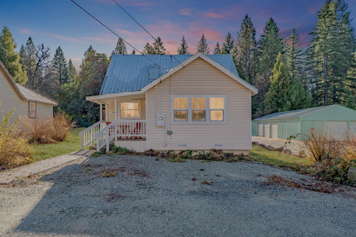 Beige house with a blue metal roof and covered porch, beside a green outbuilding among trees at dusk.