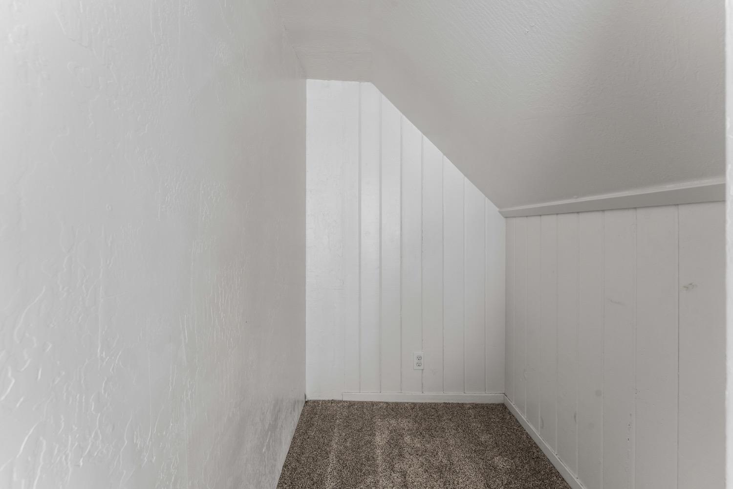 Small empty carpeted room with white paneled walls and a low sloping ceiling.