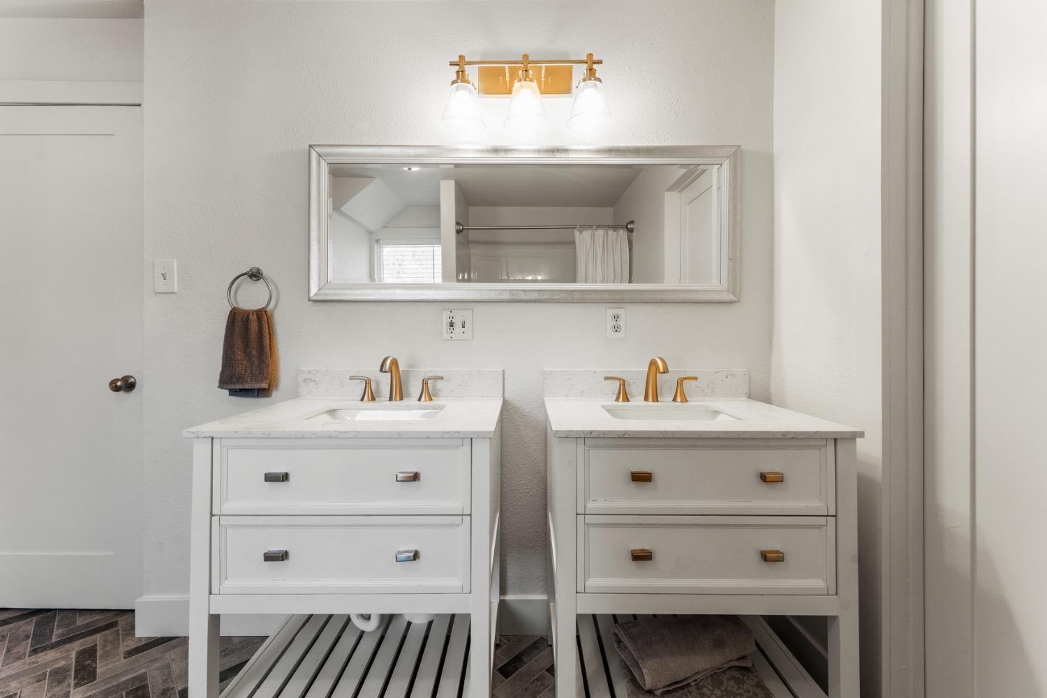 Bathroom with twin white vanities, gold faucets, a wide mirror, and dark patterned tile flooring.