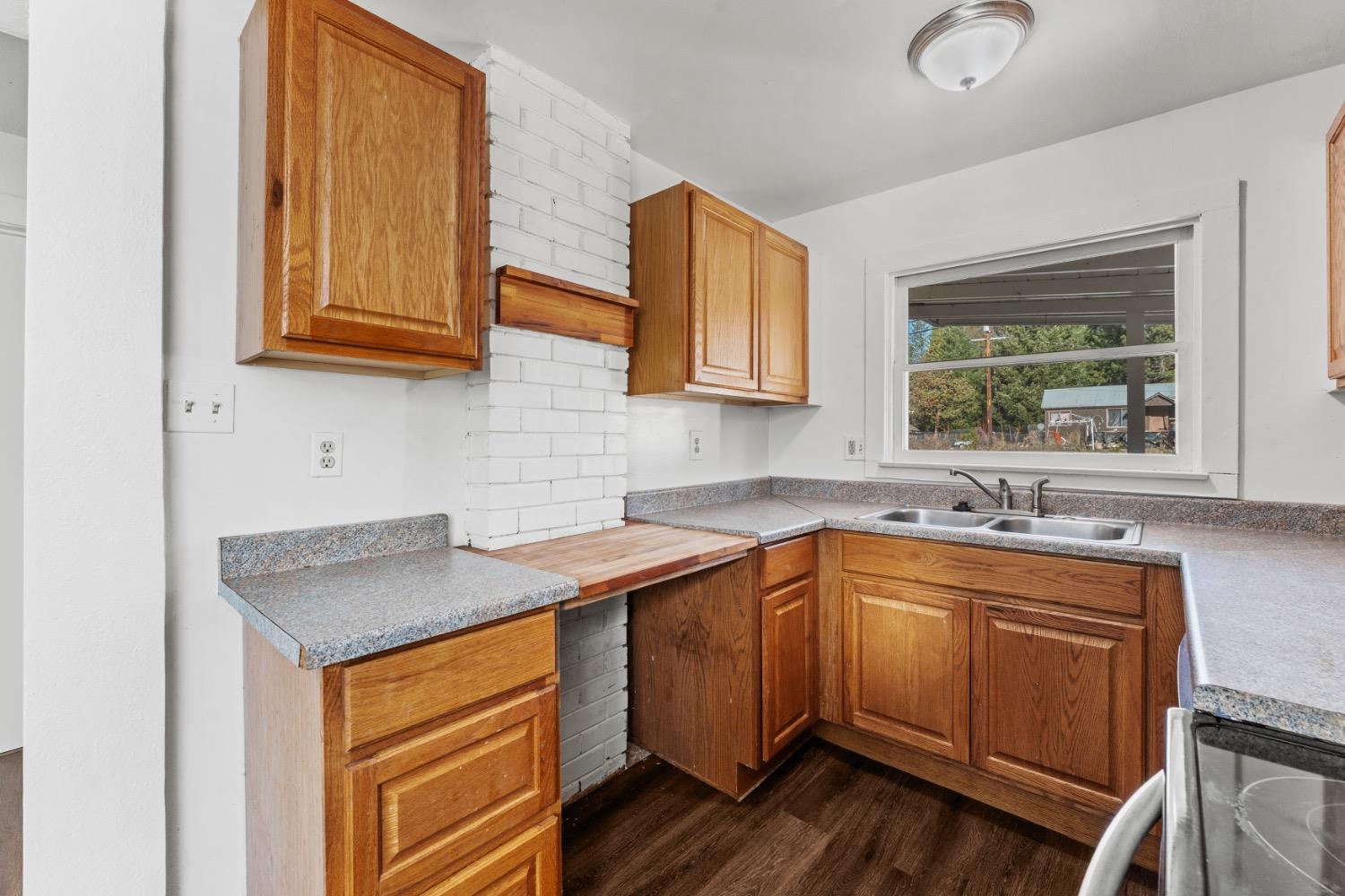 Kitchen with honey-colored cabinets, white brick wall, gray countertops, and a double-basin sink beneath a window.