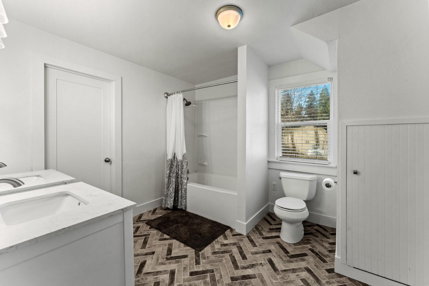 Bathroom with a white vanity, tub and patterned shower curtain, toilet beneath a window, and herringbone tile floor.