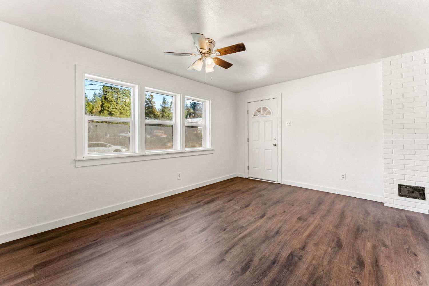 Empty room with dark wood-look flooring, three windows, ceiling fan, and white brick fireplace.