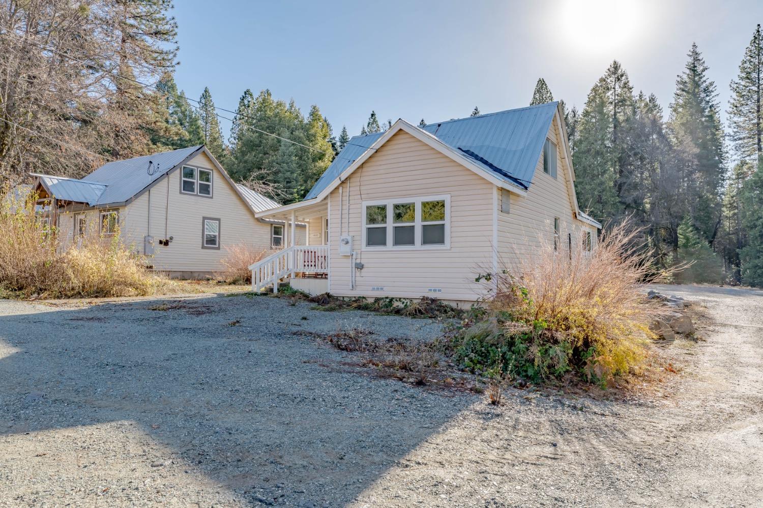 Light-colored home with a blue metal roof and covered porch beside a gravel driveway and woodland.