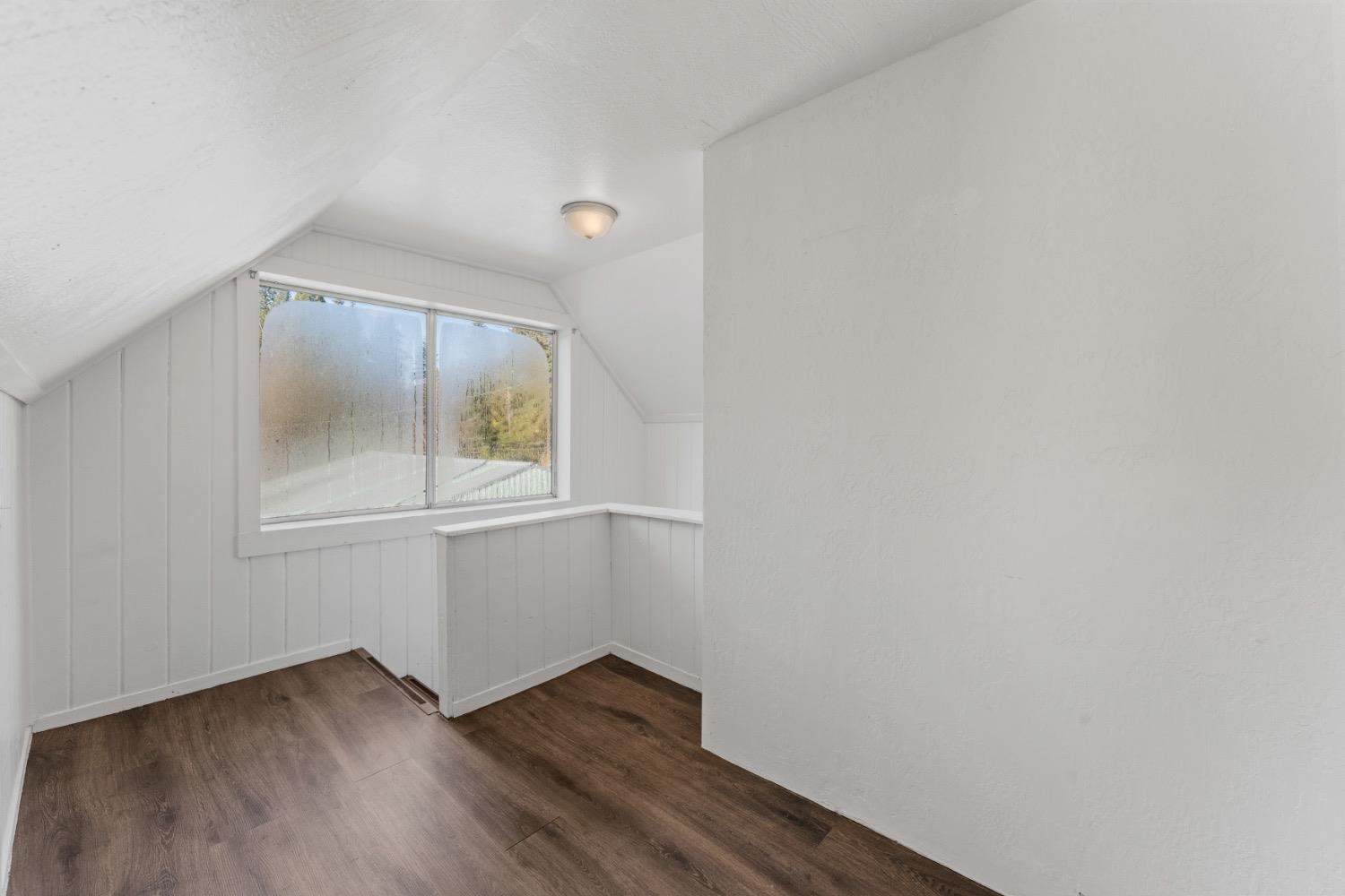 Empty finished attic room with sloped ceilings, white paneled walls, and dark wood-look flooring.