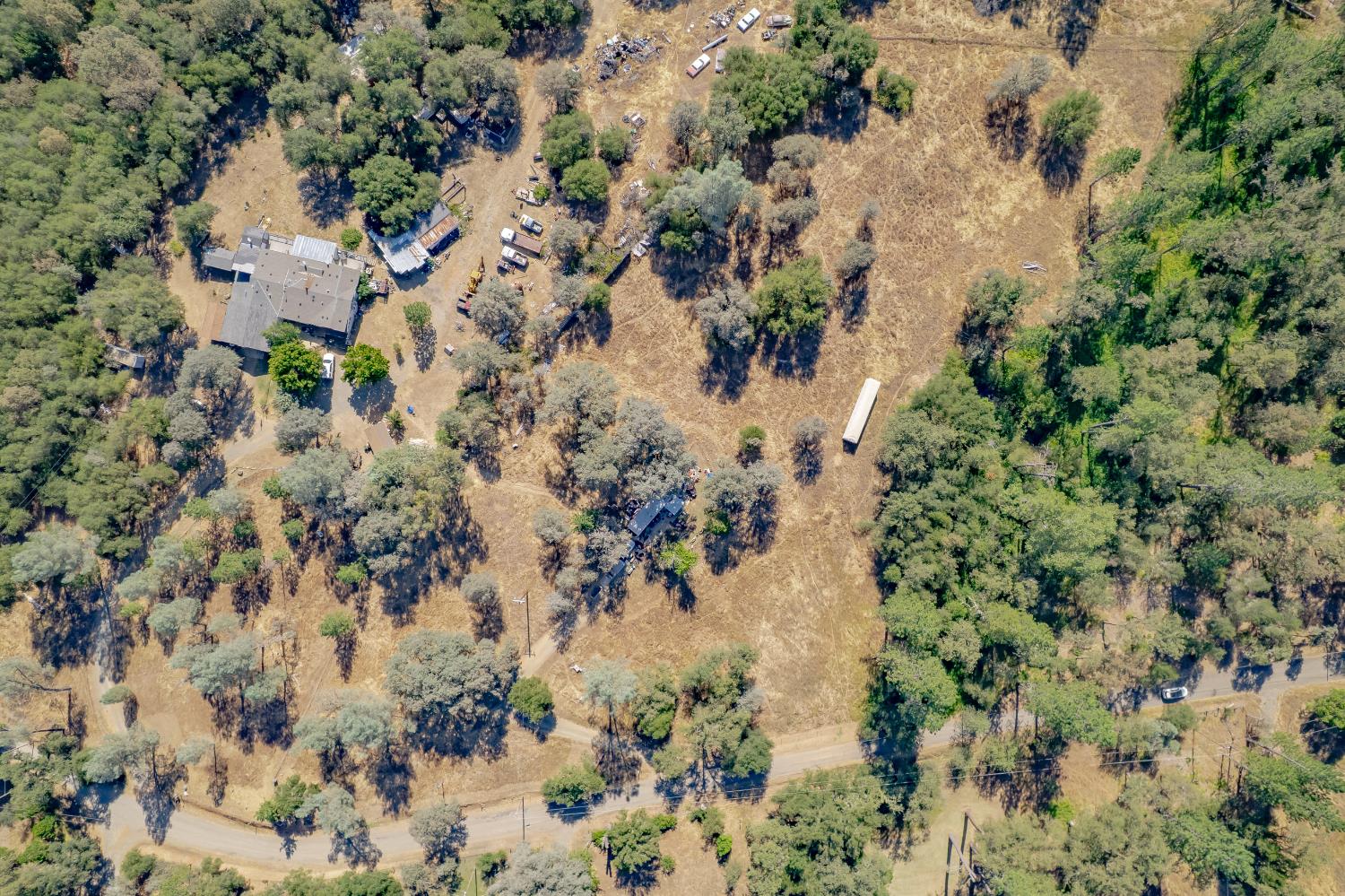 Aerial view of a rural home and outbuildings amid wooded acreage and dry open fields.