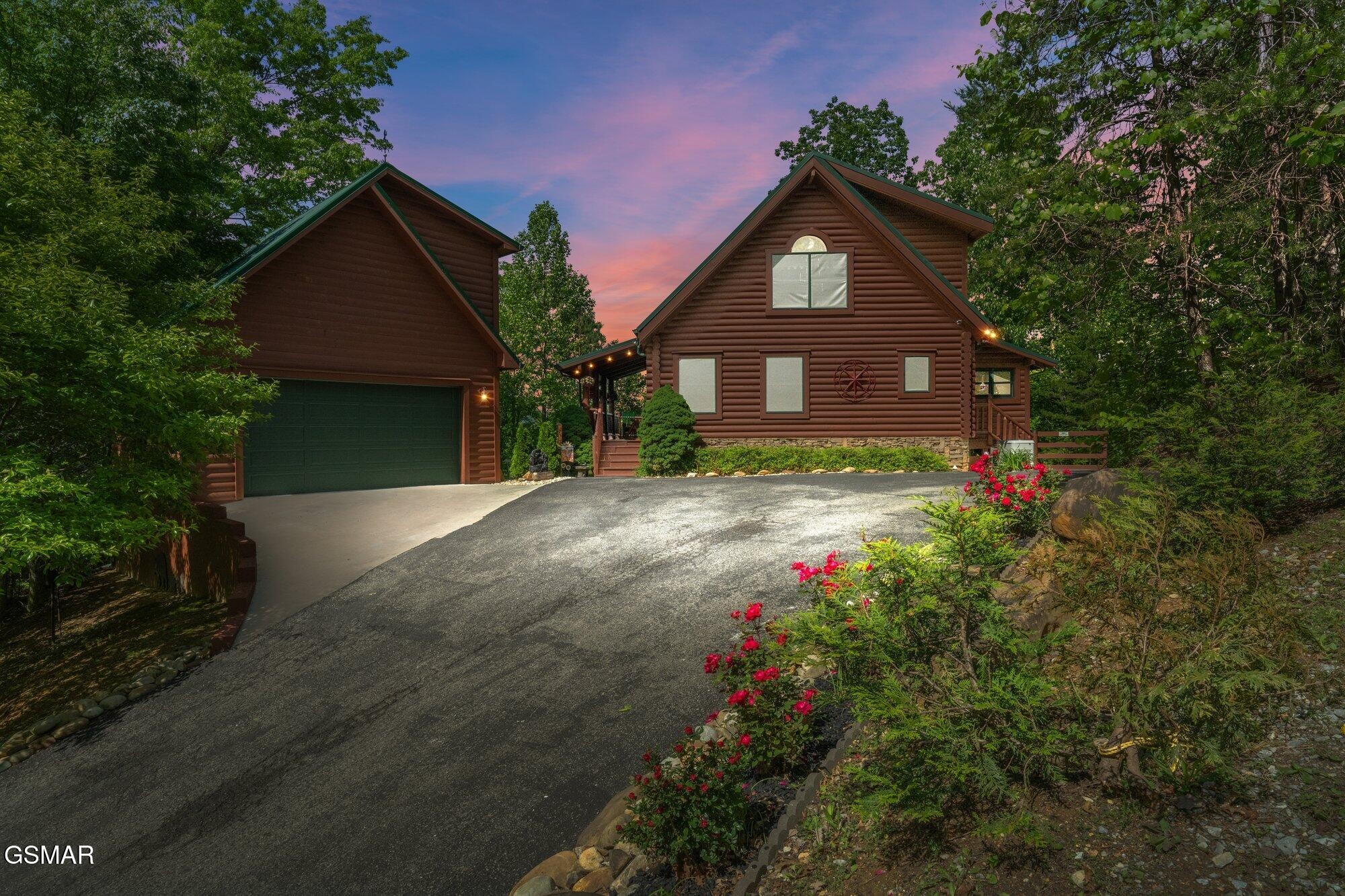 Log cabin and detached garage framed by trees and flowering shrubs at dusk.
