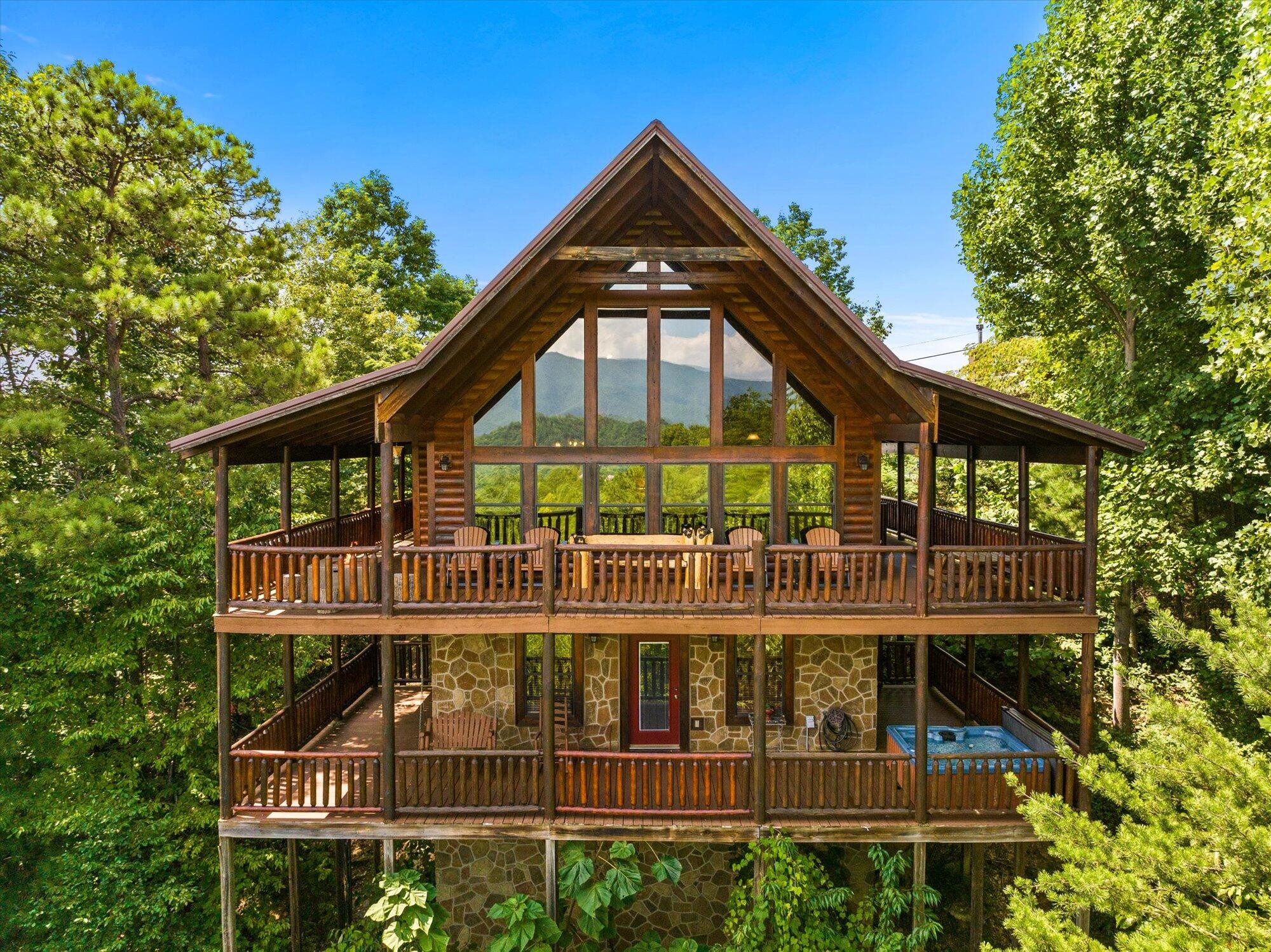 Log cabin with tiered wooden decks, stone lower level, and a hot tub among the trees.