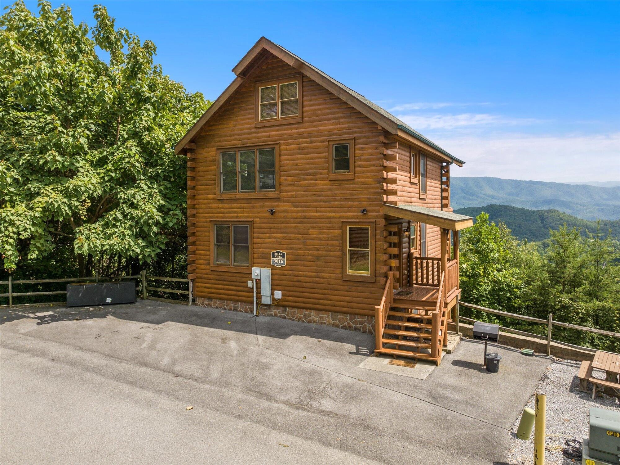 Multi-story log cabin with a wooden porch and mountain views beyond the trees.