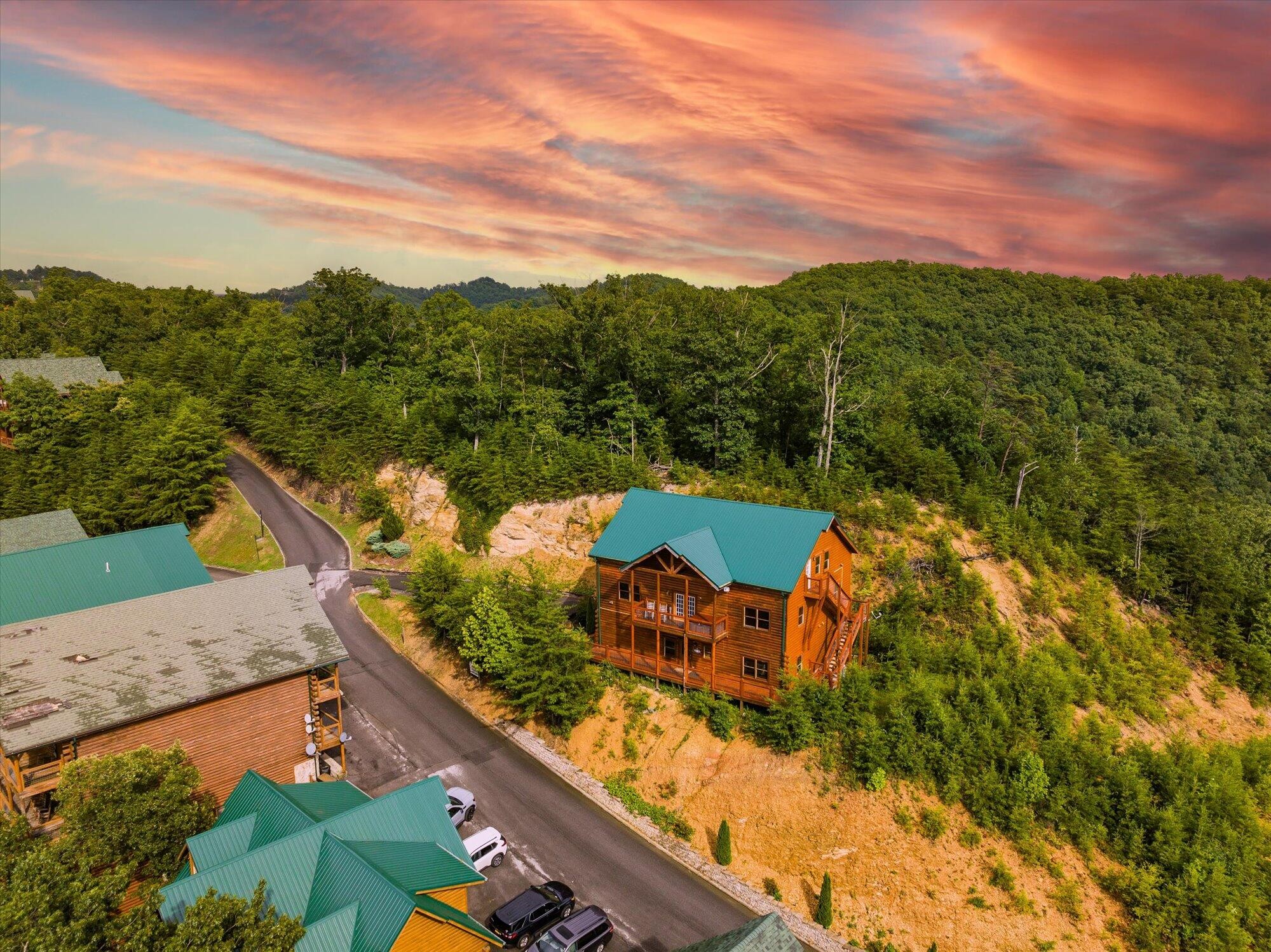 Aerial view of log cabins on a wooded hillside beneath a colorful sunset sky.